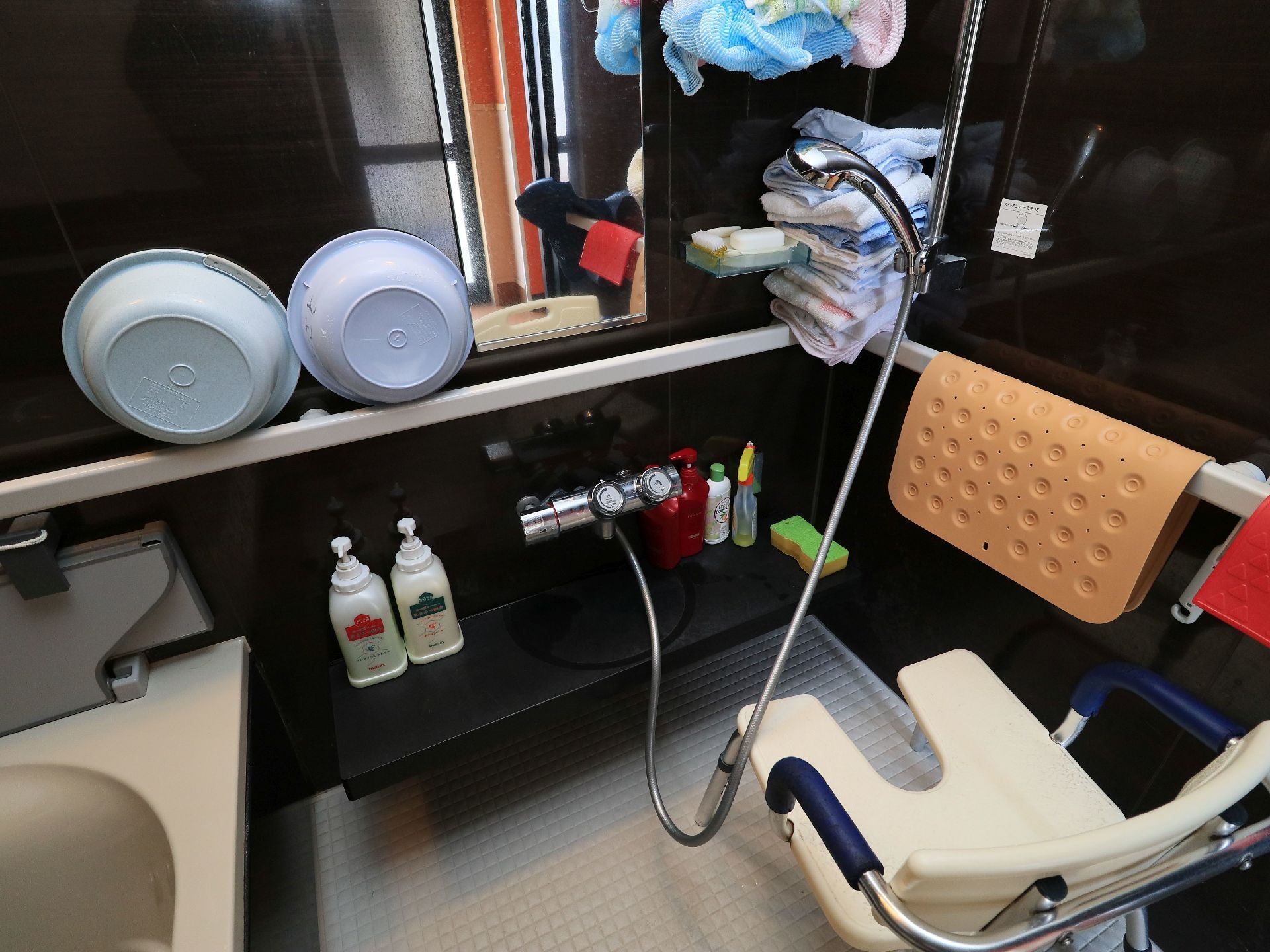 浴室の壁面には、白い手すりが渡してある。手すりにはマットや洗面器をかけている。鏡の近くに銀色のシャワーとシャワーチェアがある。