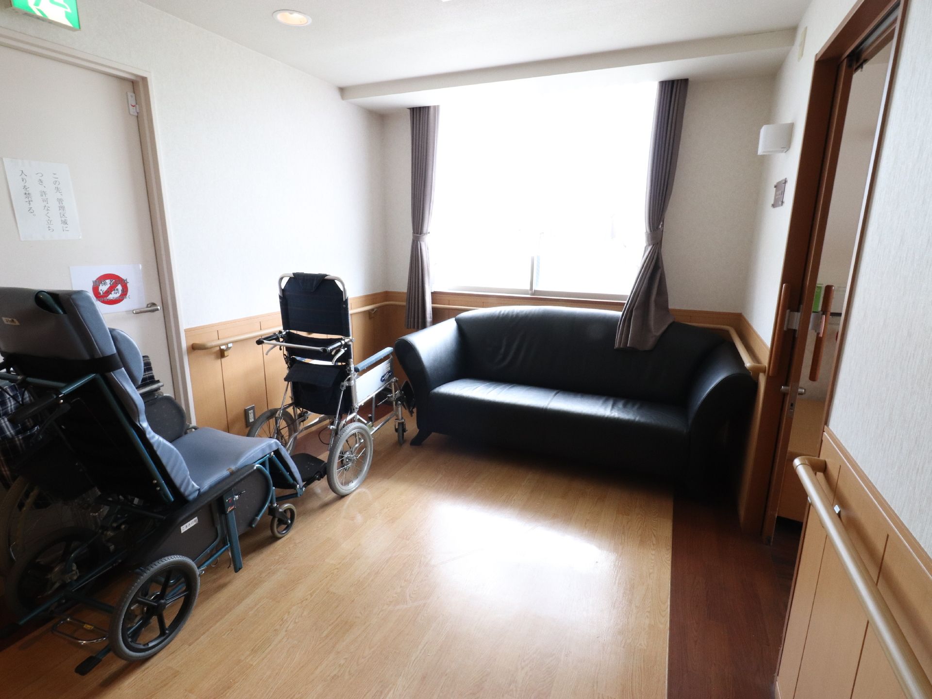 左手に非常口のある部屋は、窓際に大きめの黒いソファーが置かれてあり、数台の車いすが壁際に用意されている。
