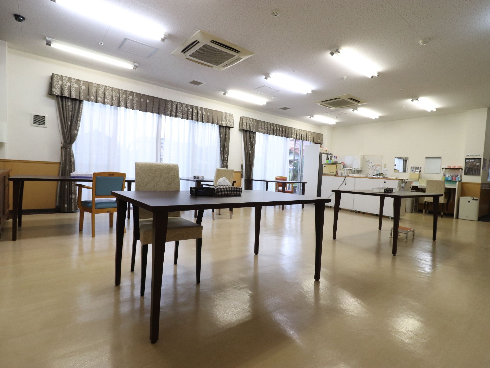 大きな窓が2つありカーテンが取り付けられている。焦げ茶色の長方形のテーブルが並んでいる。種類の違うイスがいくつか置かれている。