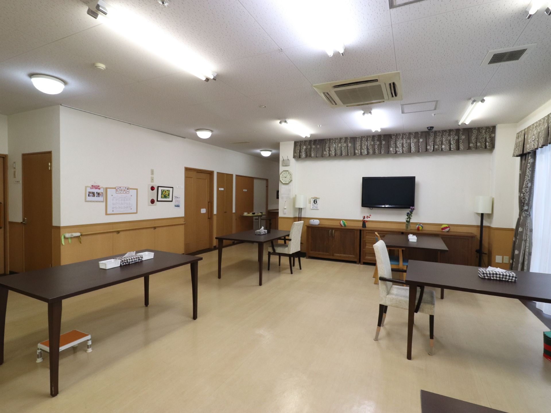 壁に手すりが取り付けられている。扉がいくつかある。テレビが設置されている。長方形のテーブルが複数並んでいる。