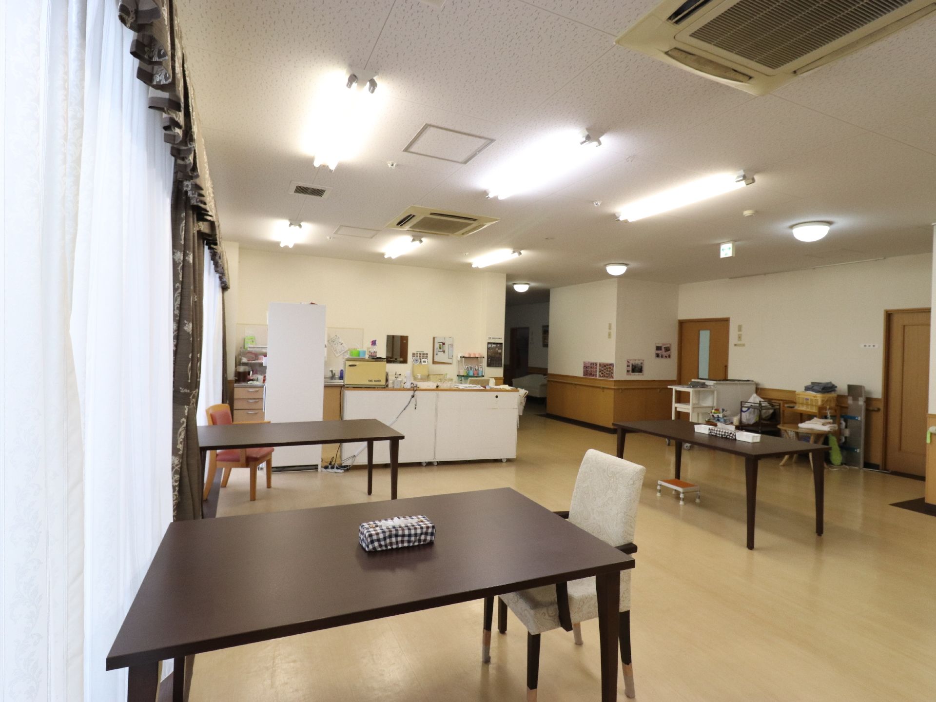 長方形のテーブルが3つある。ティッシュケースが置いてある。窓には2種類のカーテンが取り付けてある。天井にはエアコンがある。