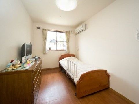 居室は落ち着いた色のフローリングと白い壁のシンプルな内装である。壁にはエアコンがあり、ベッドと家具が配置されている。