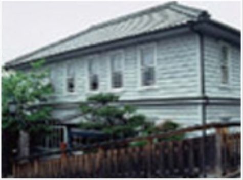 瓦屋根に白い壁の２階建ての西洋風の建物である。手前を木造塀が囲んでおり、中には樹木が何本か植えられている。