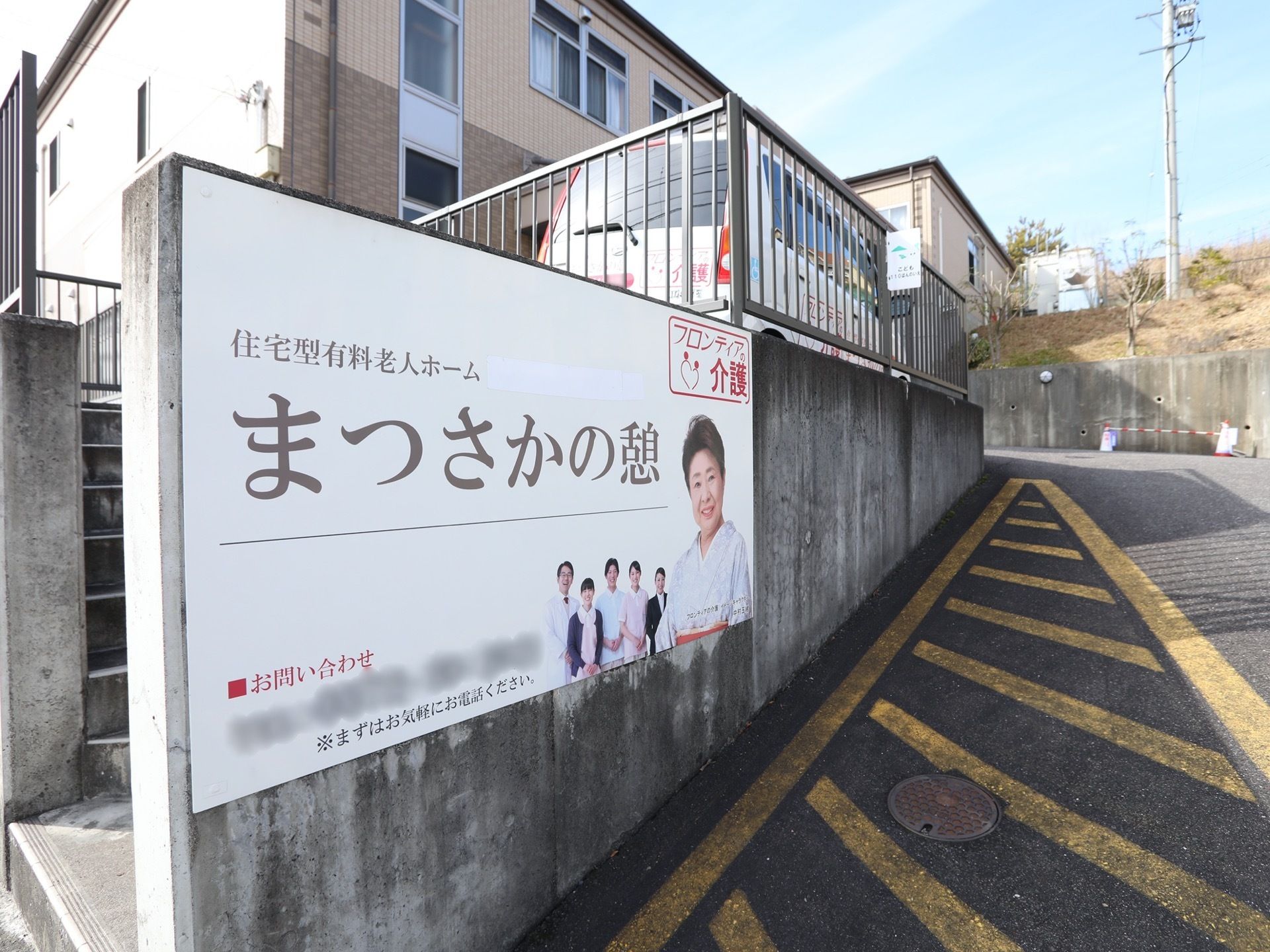 建物は道路から一段高いところにある。道路側の塀には大きな施設の看板がある。駐車場に専用車が停まっている。