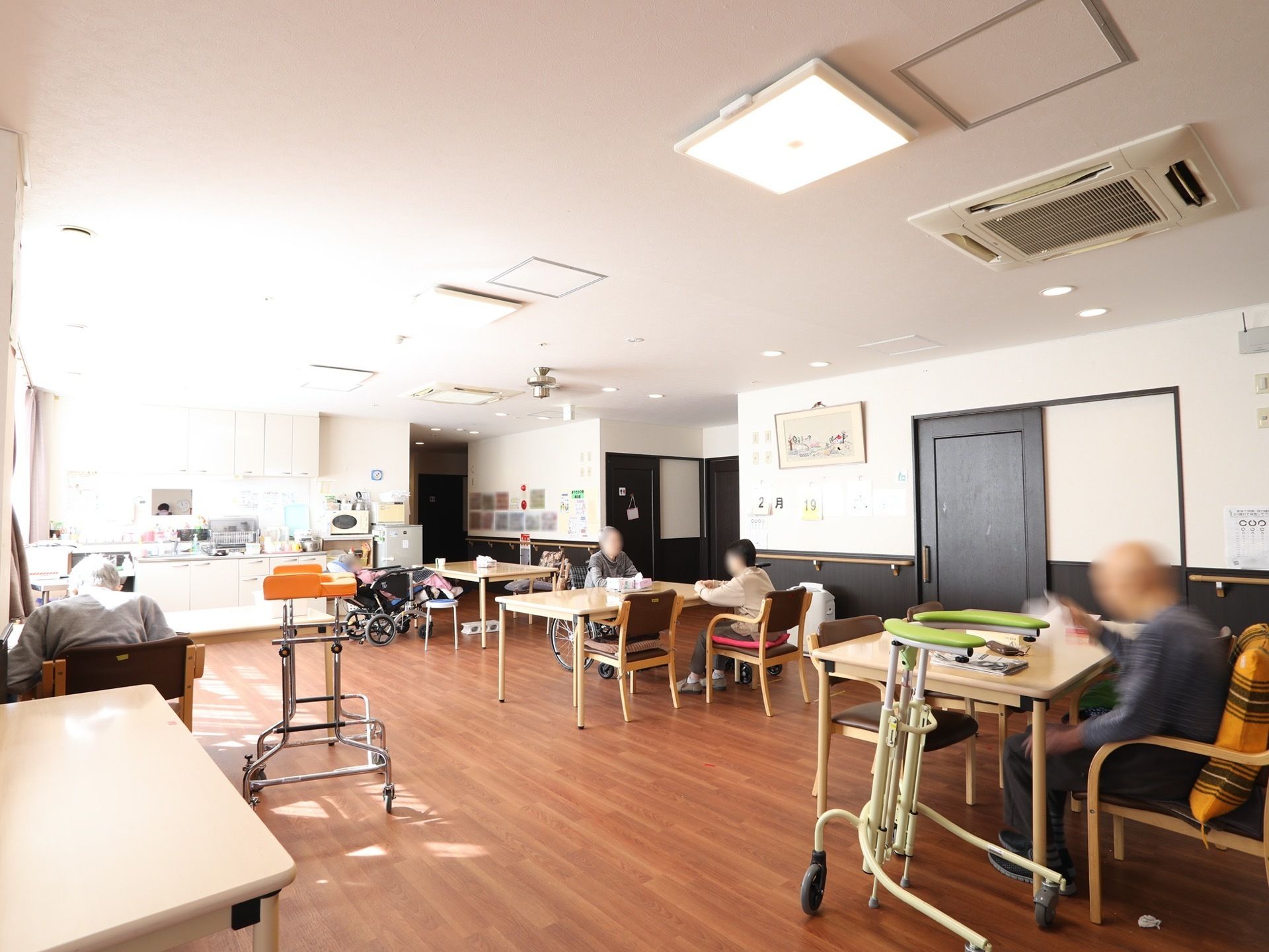 日が差し込み明るい食堂で、入居者様がくつろいでいる。歩行器を傍らに置いている方、車椅子をご利用の方もいらっしゃる。