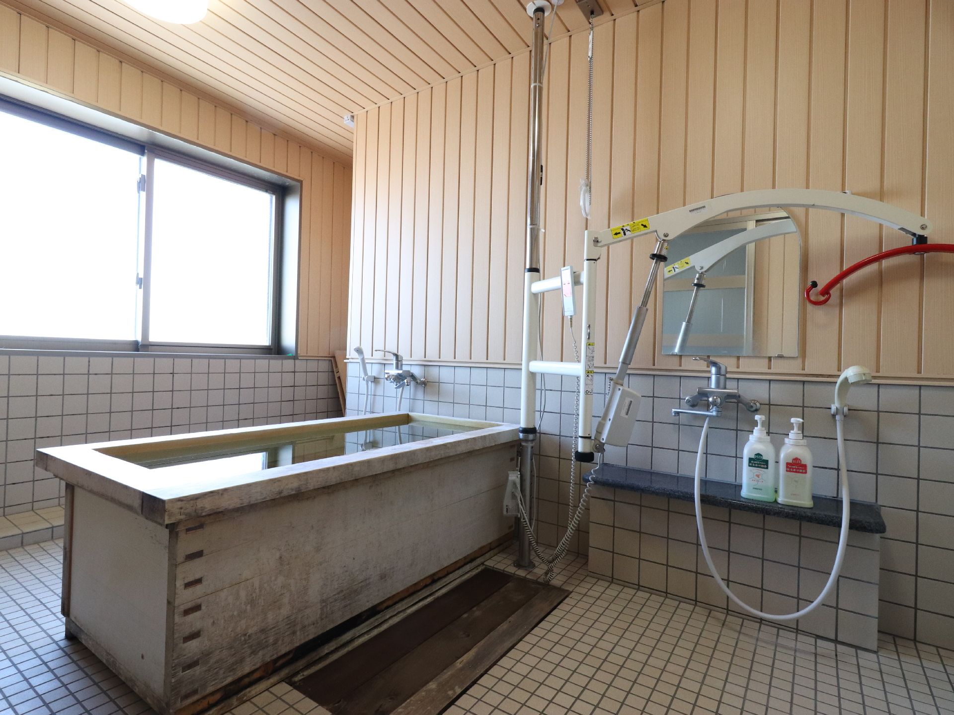 浴槽は木の造りとなっている。介助用の機械も置かれていて床や壁の下半分はタイルを使っている。シャワーが二つ付けられている。