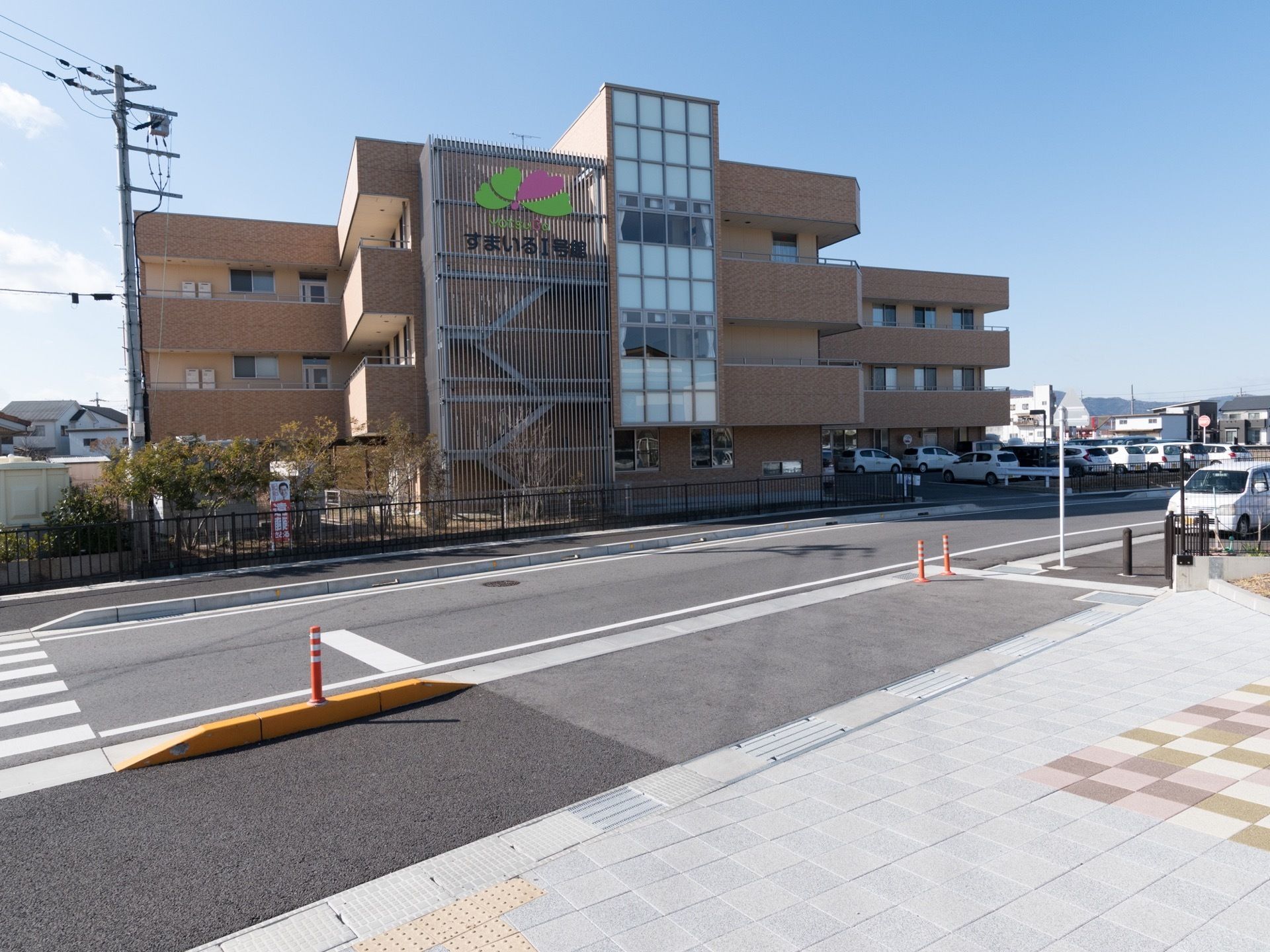 施設は鉄骨造り３階建ての建物で、外壁はベージュ色のタイルが貼られている。外階段のフェンスに施設名が掲示されている。