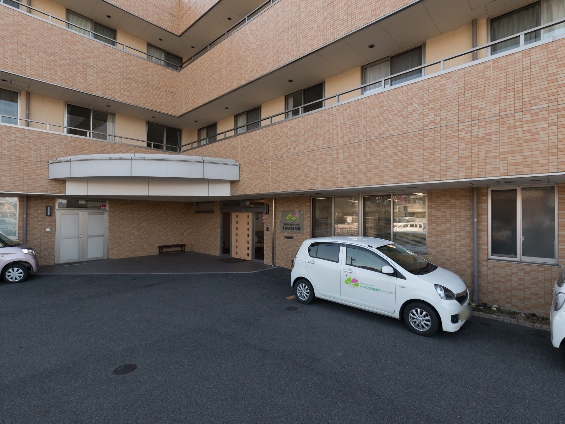 施設の外壁やベランダ壁には、ベージュ系のタイル張りである。玄関前にはポーチがある。施設名が表示された看板が貼られている。