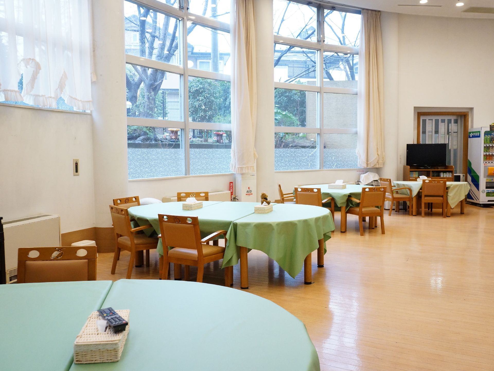 高い天井に壁面は窓が連なり、明るく解放感たっぷりな食堂。車いすのまま食卓につけるテーブルの高さ。テレビもあり、皆が集える場所。
